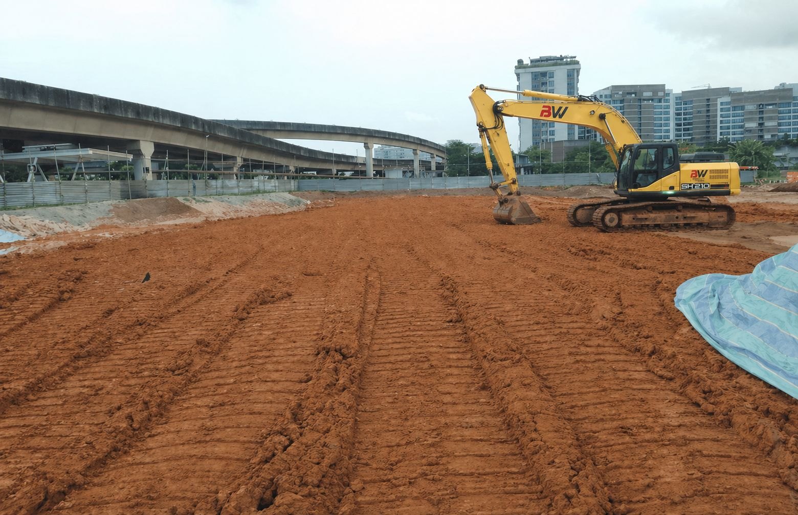 Buildwick BW-branded SH210 excavator on freshly graded earthworks site beside an elevated MRT viaduct