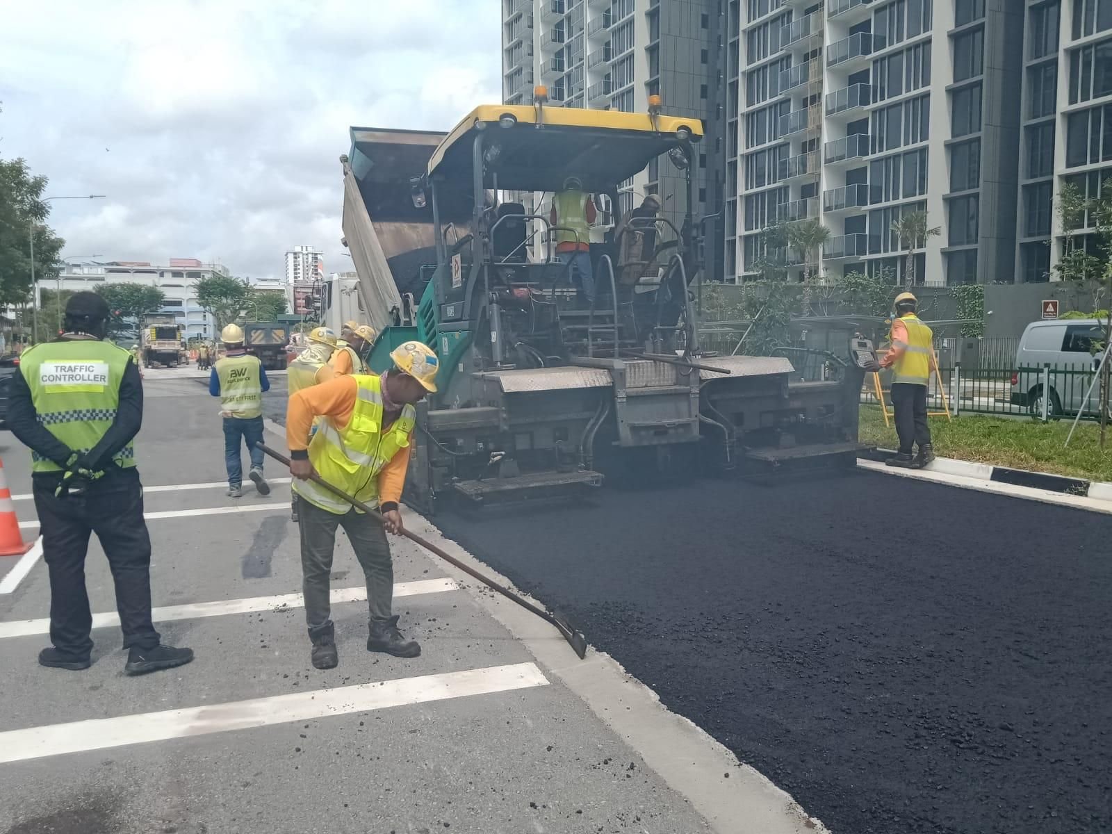 Buildwick crew laying fresh asphalt with paver machine alongside live traffic — high-vis PPE, traffic controller and cones in place